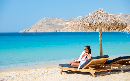 women at Mykonos beach during summer with umbrella and luxury beach chairs beds, blue ocean with the mountain at Elia beach Mikonos Greece.