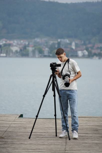 A vertical of a Caucasian guy on a wooden pier while shooting with a professional camera on a tripod