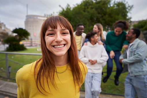 Portrait of a Young red-haired girl with wide smile and piercing look at the camera. Group of friends gathered in the background of the image. Colleagues hanging out in the city. Outdoors.