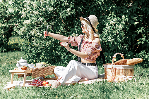 Portrait of a young blonde woman in a hat and sunglasses at a picnic in the park .Takes a selfie on a smartphone on a summer vacation.Video chat.