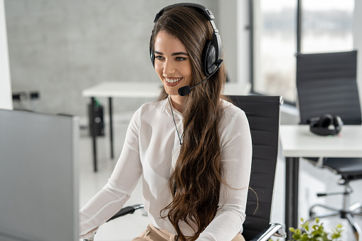 Friendly female call center operator working in office. Young beautiful woman in modern office working as call center agent with headset on head.