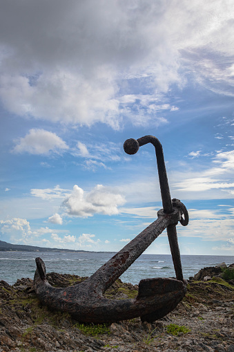 A vertical closeup of an old rusty anchor on the shore against the background of the sea and sky.