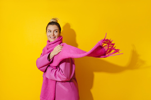 Portrait of young cheerful woman wearing pink coat and wool scarf against yellow background