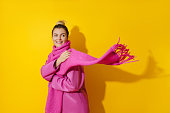 Young cheerful woman wearing pink coat and wool scarf against yellow background