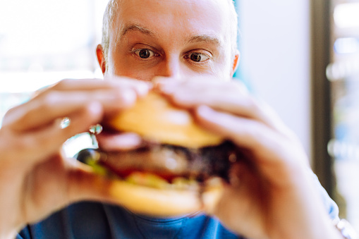 Close up image depicting a man holding a fresh burger loaded with beef and crispy bacon and salad. He is sitting in the restaurant and his eyes are wide with amazement at the size of the burger. Room for copy space.