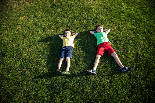 Picture of two brother having fun in the park, two cheerful children laying down on green grass, little boy and his friend playing outdoors, best friends, happy family, love and happiness concept.