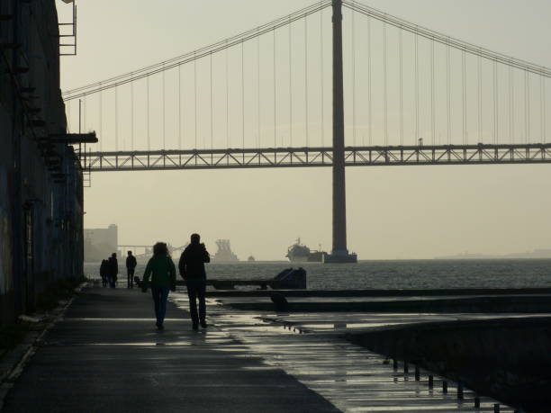 backlit people walking in Cacilhas by the Tagus river with the 25 de Abril bridges in the background, Portugal
