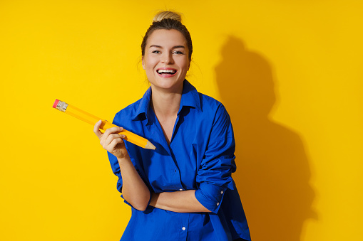 Cheerful young woman wearing blue shirt holding giant pencil on yellow background