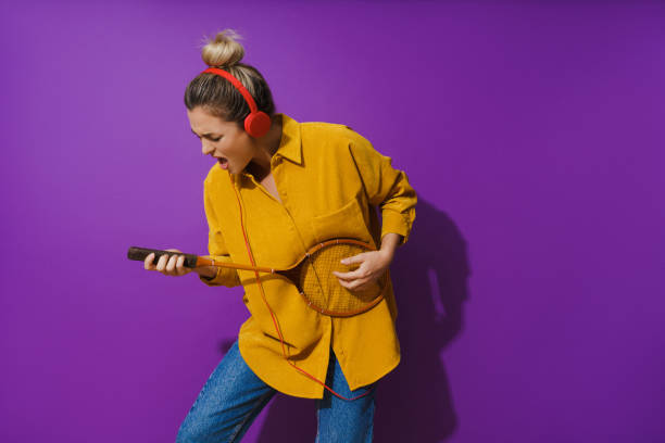 Cheerful girl is jamming out in her yellow shirt while wearing red headphones. She's strumming away on her racket like it's a guitar. Cheerful girl is jamming out in her yellow shirt while wearing red headphones. She's strumming away on her racket like it's a guitar, against a vibrant purple background. air guitar stock pictures, royalty-free photos & images