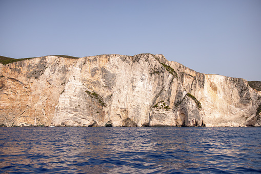Amazing view from the boat of the high steep cliffs in Greece on a beautiful sunny summer day. The water is calm and blue. The cliffs are illuminated by the sunlight.