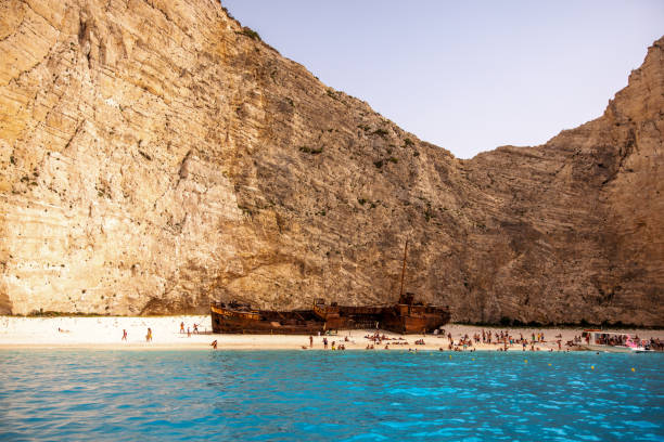 Beautiful Navagio beach in Greece, Zakynthos with a shipwreck. The water is blue and clear. The beach is surrounded by amazing steep cliffs. The beach is full of tourists admiring the shipwreck. The weather is sunny and the sun is shining.