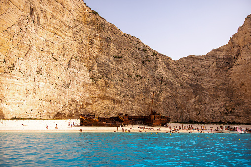 Beautiful Navagio beach in Greece, Zakynthos with a shipwreck. The water is blue and clear. The beach is surrounded by amazing steep cliffs. The beach is full of tourists admiring the shipwreck. The weather is sunny and the sun is shining.