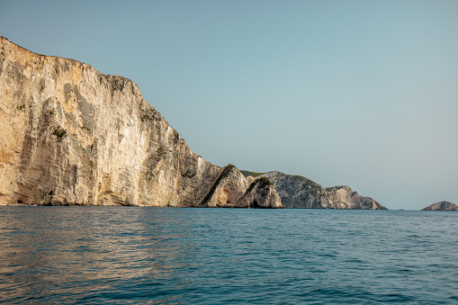 Amazing view from the boat of the Greek coastline and the calm blue sea. The sun is shining and illuminating the walls of the cliffs. The sky is clear and blue.