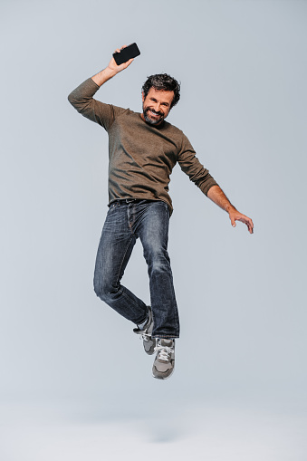 Studio shot of an happy screaming and exited mature man with casual clothings jumping high with his mobile phone in the hand on grey background. Part of a series with similar poses and different facial expressions.