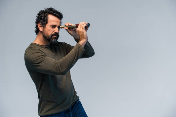 Studio shot of a casual dressed mature man with natural black and gray hair looking into the distance with binoculars on grey background in direction of the copy space stock photo