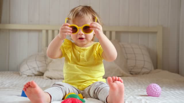 Child playing with colorful plastic toys on beige cozy bed in sunny white bedroom. Educational game for baby and toddler. Young girl play indoors. Wearing sunglasses. Kid preparing to vacation, travel