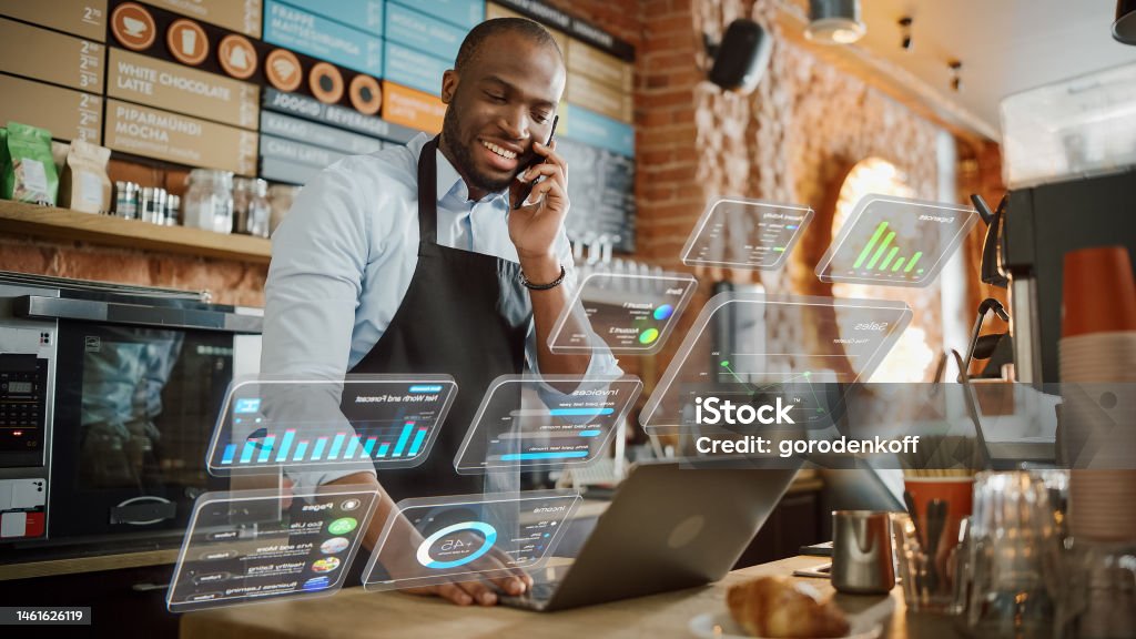 African American Coffee Shop Owner is Working on Computer and Talking on a Phone in a Cozy Cafe. Augmented Reality Icons Popping Out of Restaurant Manager's Laptop: Small Black Business Concept. Delicatessen Stock Photo