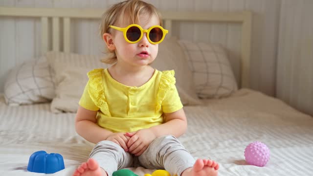 Child playing with colorful plastic toys on beige cozy bed in sunny white bedroom. Educational game for baby and toddler. Young girl play indoors. Wearing sunglasses. Kid preparing to vacation, travel