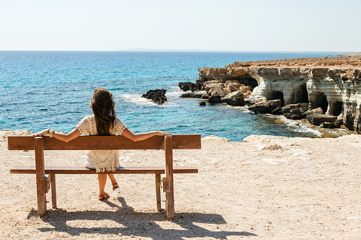 Young woman sits on a bench near sea cliffs at Cape Greco viewpoint, enjoying the scenery and natural beauty.