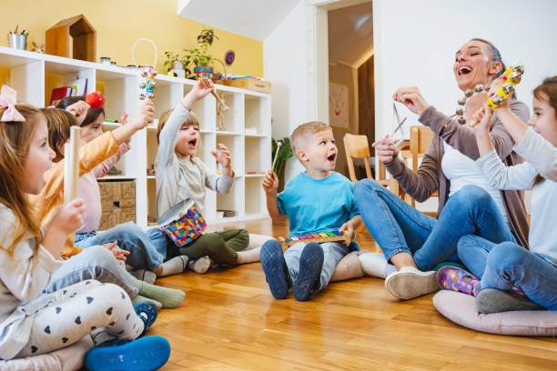 Kindergarten teacher with children sitting on the floor having music class, using various instruments and percussion. Teacher with children in a Kindergarten Classroom. Healthy learning environment. Kindergarten teacher, building relationships with the kids. Cheerful preschool children preschool classroom jobs stock pictures, royalty-free photos & images