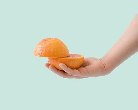 Woman hand holds two halves of a red grapefruit in her hand on a turquoise, green, blue background. Summer healty fruit concept.