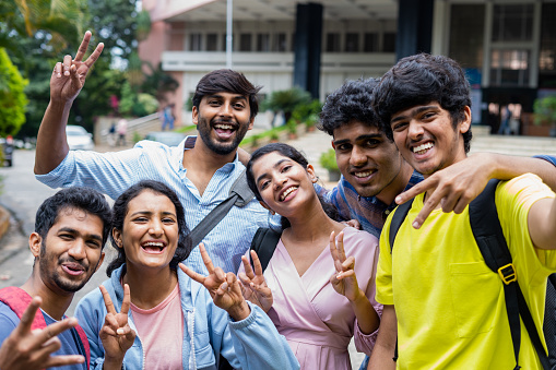 Group of smiling college students taking selfie by holding camera at college campus - concept of friendship, education and success