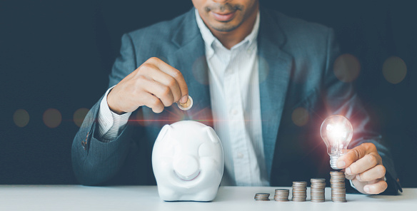 Businessman hand holding lightbulb putting a coin into the piggy bank, saving energy, accounting finance in home and family concept, saving world and energy power ideas for saving money concept.