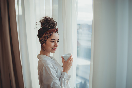 Young business woman, dressed in a white shirt, relaxes in the morning before all business obligations with coffee and looking out the window of the hotel room