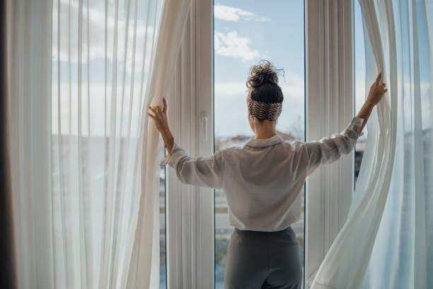A young woman opens the curtains in the room in the morning at sunrise stock photo