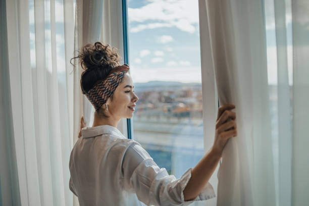 one young woman opens the curtains in the room in the morning at sunrise, she is dressed in a white shirt, ready for work and daily duties, morning routine - sunny apartment window sky imagens e fotografias de stock