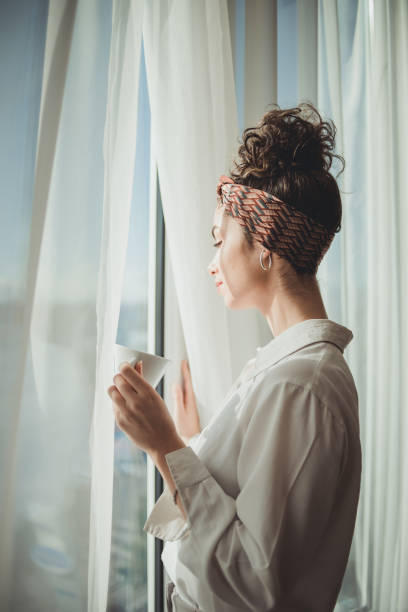 One young woman opens the curtains in the room in the morning at sunrise, she is dressed in a white shirt, ready for work and daily duties, morning routine stock photo
