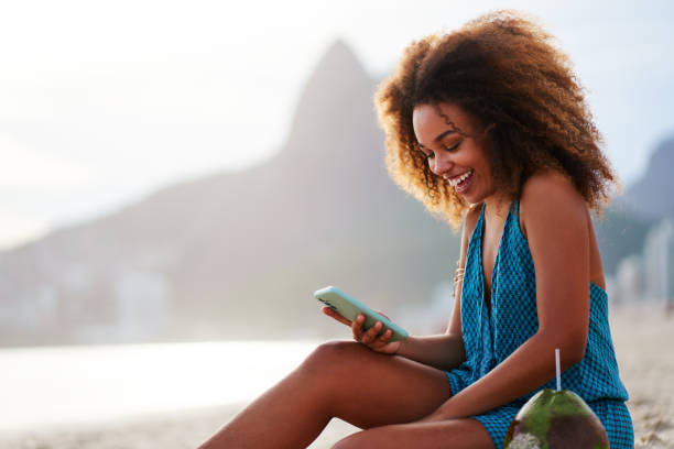portrait smiling happy young brazilian woman sitting on the beach holding and looking at cell phone in Ipanema stock photo