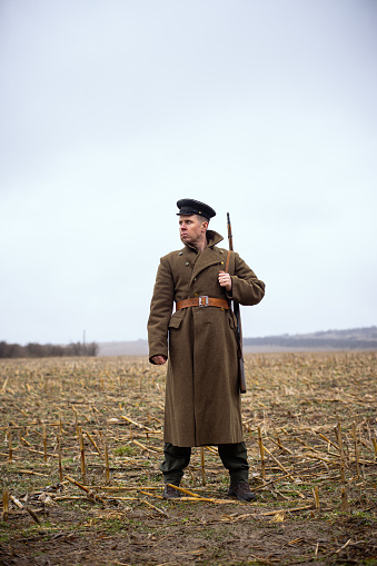 a retro soldier in an old coat with a weapon on his shoulder stands in a field and looks to the side. USSR soldier of the First World War