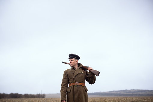 a retro soldier in an old coat with a weapon on his shoulder stands in a field against the background of the sky. USSR soldier of the First World War.