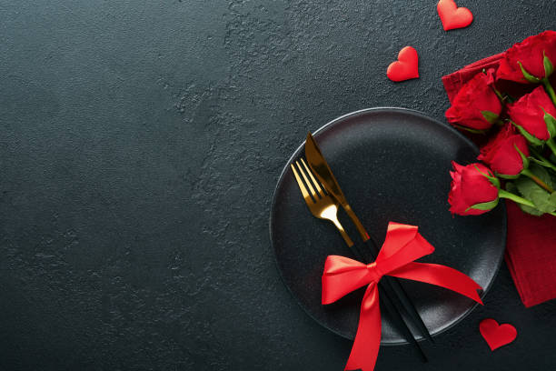 istock Valentines day romantic table setting. Empty closeup of red roses, wine, candles, dinner black plate, knife, fork and decorative silk hearts on black background. Holiday concept. Copy space. Top view 1461650989