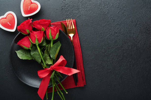 istock Valentines day romantic table setting. Empty closeup of red roses, wine, candles, dinner black plate, knife, fork and decorative silk hearts on black background. Holiday concept. Copy space. Top view 1461651053
