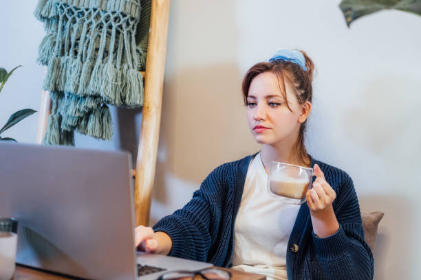 ritratto di giovane donna concentrata che beve caffè mentre usa il computer portatile. accogliente spazio di lavoro nella moderna casa interna scandi con piante verdi. lavoro a distanza a casa. istruzione o intrattenimento online. - topics selective focus concepts blog foto e immagini stock