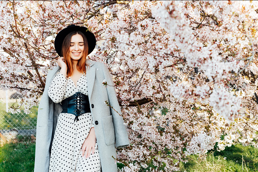 Laughing romantic woman in the blooming garden. Springtime. Tenderness young model with closed eyes in a gray coat and hat. Generation z, millennials. Sensitive mood. Selective focus, copy space.