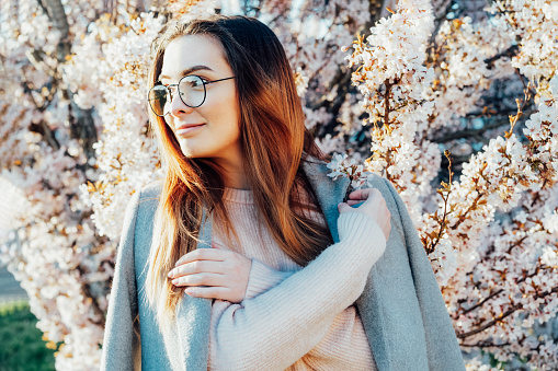 Young caucasian brunette fashion woman in eyeglasses, gray coat and pink clothes near blooming tree with sunset light. Elegant street fashion. Generation Z girl enjoy spring mood. Selective focus
