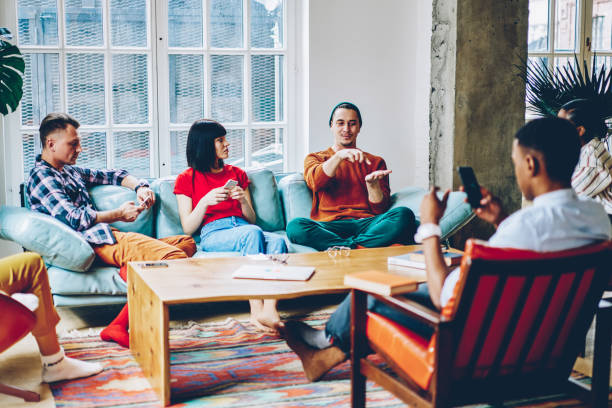 Young content male in casual clothes explaining pointing at hand playing game with friends spending time sitting on sofa in flat