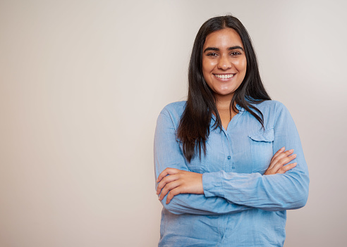 Young Indian woman stands arms folded beige wall, blue shirt office attire. High quality photo