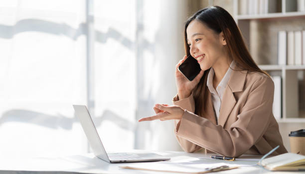 Attractive smiling asian businesswoman in light brown shirt talking on the phone with customer to name sales and checking finances on graph papers and laptop in office stock photo