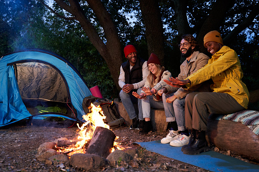 Young multiethnic group of friends camping outdoors and sitting around campfire on the forest in the evening, speaking and enjoying their trip adventure. Copy space.