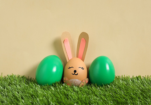 Bright eggs and brown one as Easter bunny on green grass against beige background