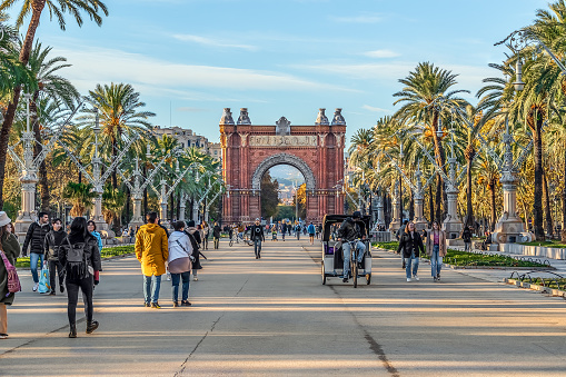 Barcelona, Spain - November 26, 2021: People walk in front of Triumphal Arch on Passeig de Lluis Companys in Barcelona. Many tourists and a tuk-tuk on the promenade between palm trees and street lamps