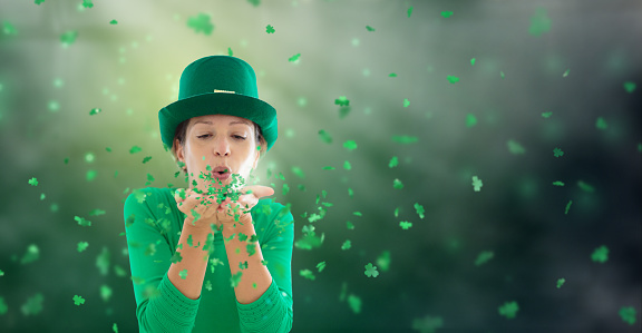 St Patrick’s day celebration. Woman in green leprechaun hat blowing shamrock confetti in Irish pub. Saint Patrick day party. Clover leaf invitation banner. Ireland tradition.