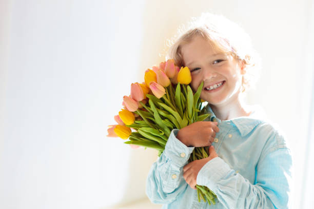 Child with flower bouquet. Mother’s day greeting. Little boy with bunch of tulips. Gift for mom. Spring birthday present. Kid in sunny living room holding dozens of flowers for mother.