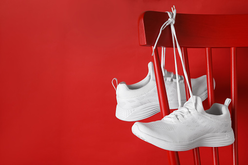 Stylish sneakers with white shoe laces hanging on chair against red background. Space for text