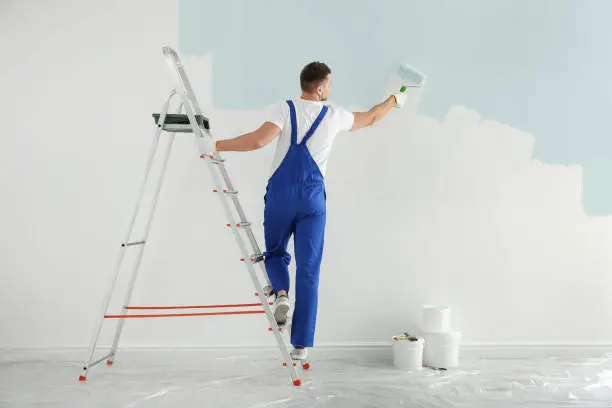 Photo of Man painting wall with light blue dye indoors, back view