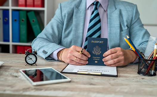 Close-up of male hands filling out visa documents at a table. On the table there is an American passport and an American flag